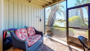 A covered outdoor seating area with a wicker sofa and chairs, a view of palm trees and buildings, and a large window. - Cozy condo w/ a sunroom plus a shared pool & tennis - minutes from the beach (Miramar Beach)