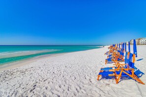 Uma praia com fileiras de cadeiras de praia azuis e laranja, e um céu azul límpido.