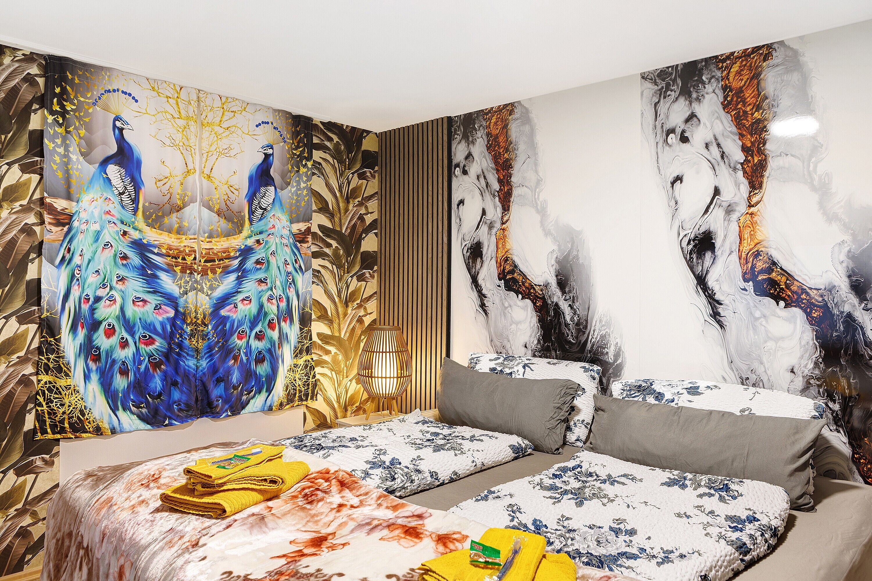 Un dormitorio con un amplio mural de pavo real, una cama con ropa de cama floral y una lámpara moderna.