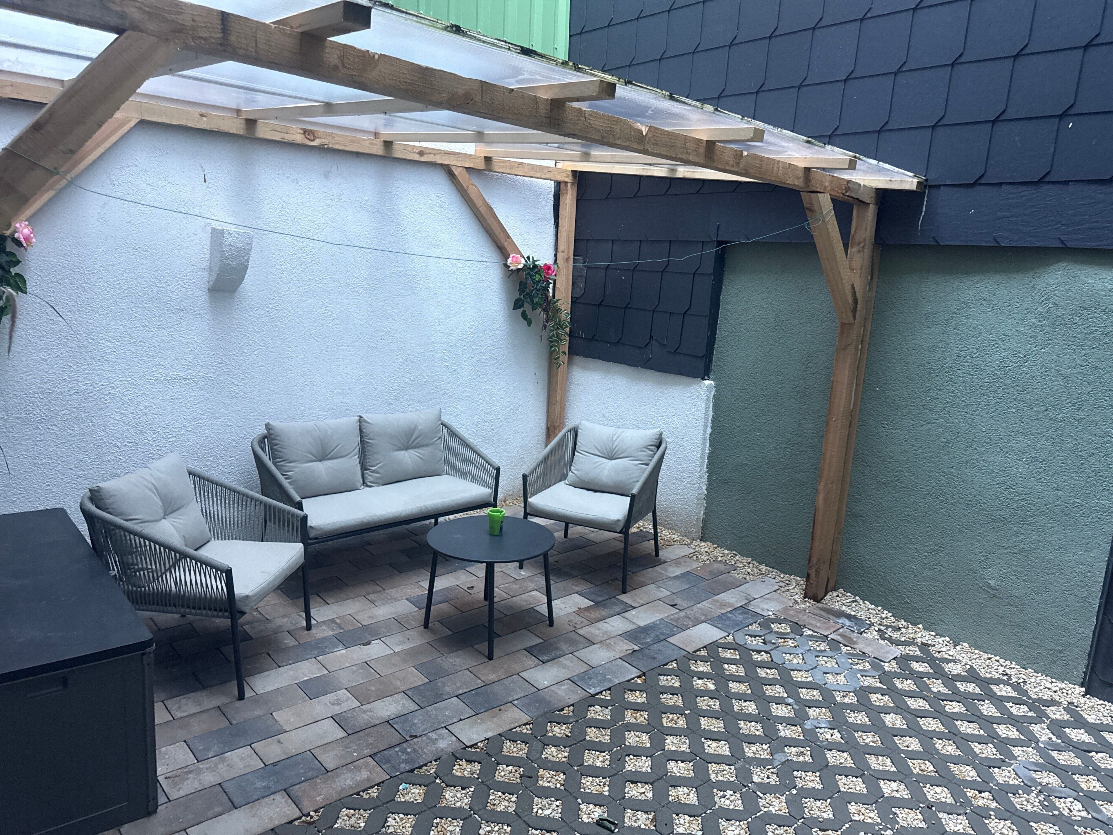 En overdækket terrasse med et træpergola, en grå sofa, to lænestole, et lille rundt bord og et sort skab.