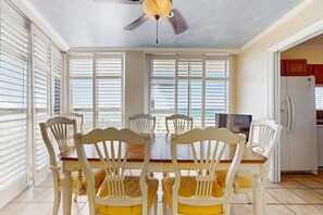 A dining area with a wooden table and white chairs, a ceiling fan, and a view through large windows. - Spacious beachfront condo with full kitchen, shared pool and beach access (Miramar Beach)