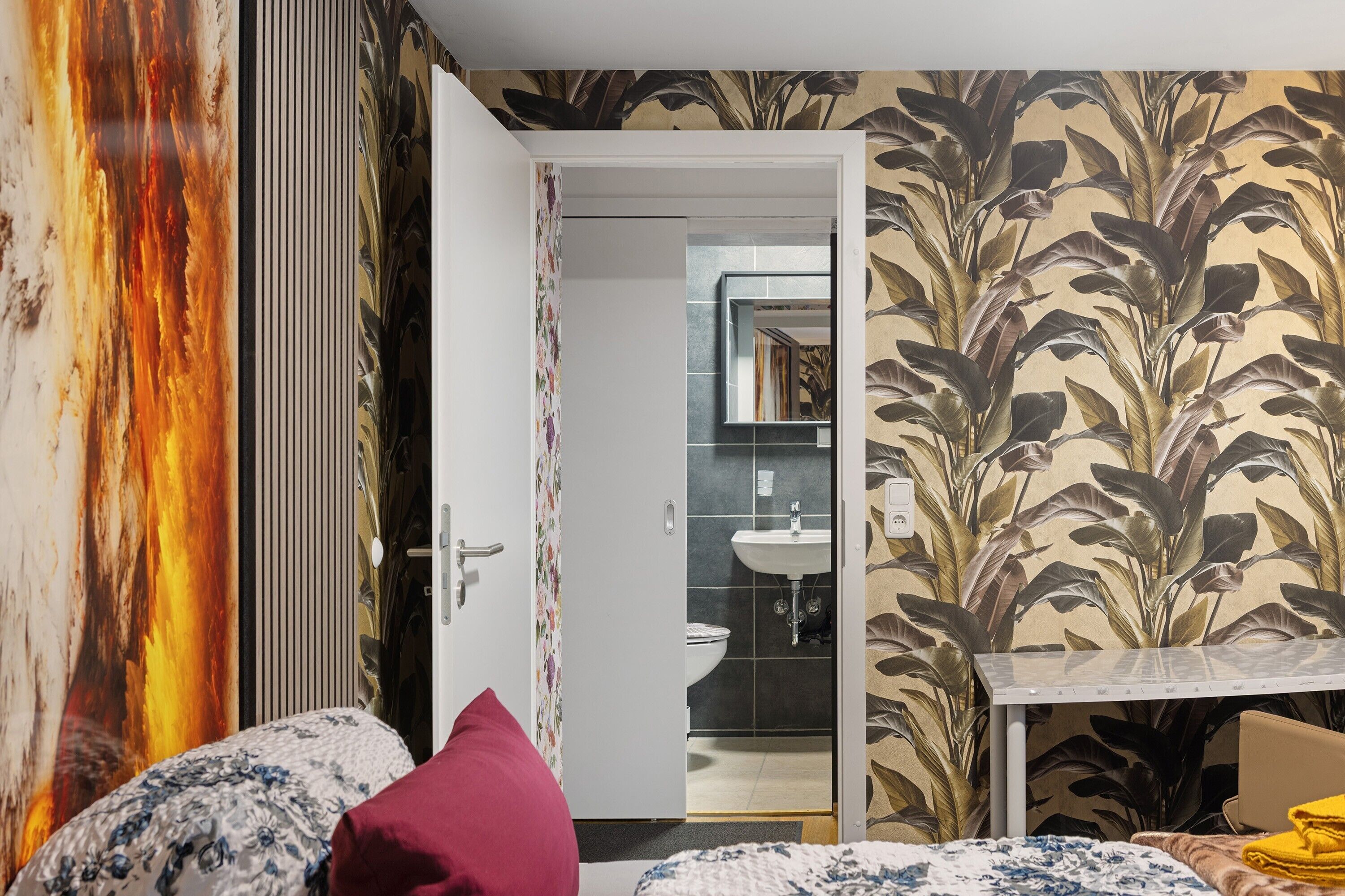 Un dormitorio con cama, mesita de noche, almohada roja, una mesa con silla, y un baño visible a través de una puerta abierta.
