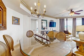 A dining area with a glass table, wicker chairs, a chandelier, and a television in the background. - Gulf view condo w/ balcony, shared pool/hot tub/tennis/gym & beach access (Miramar Beach)