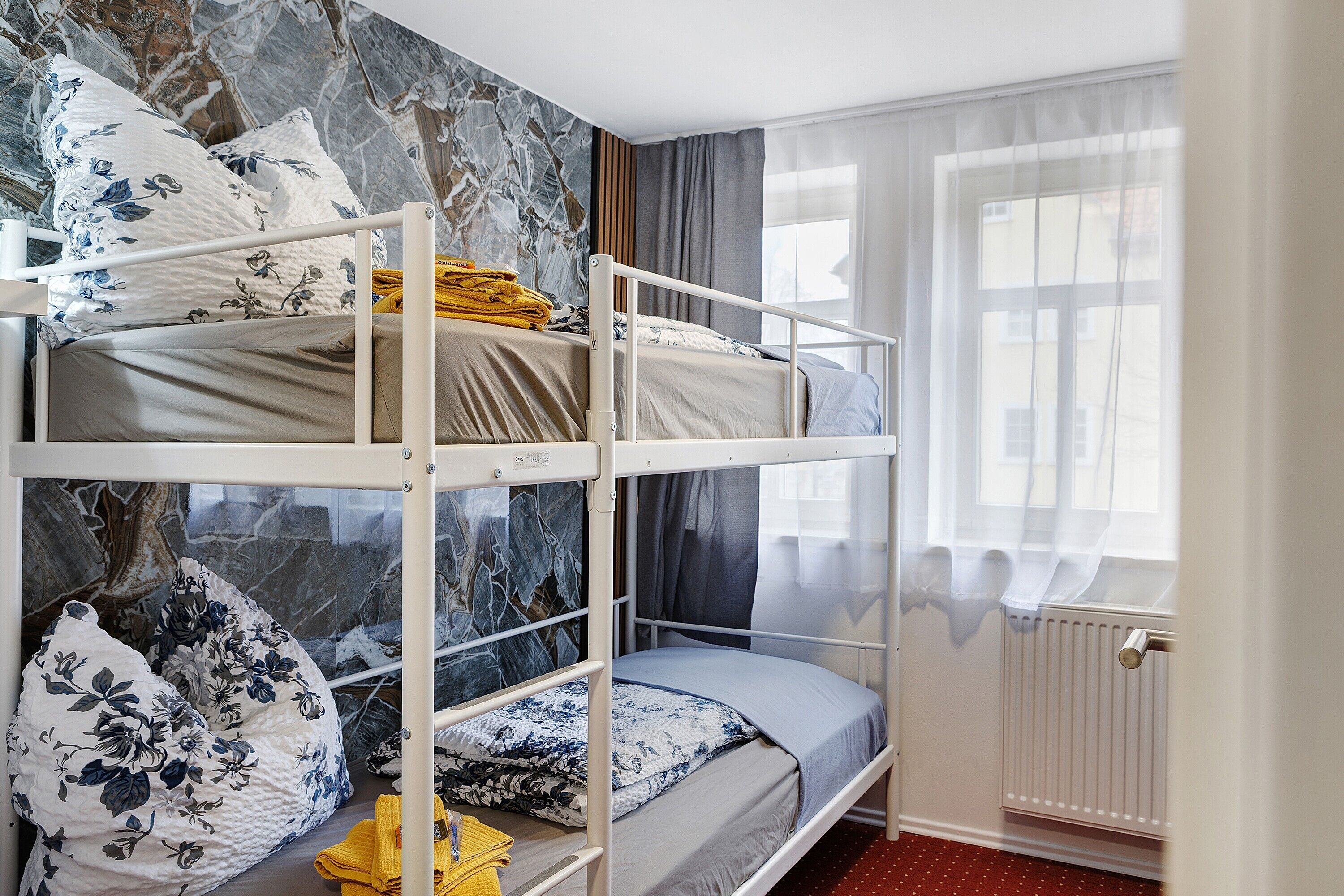 Een slaapkamer met een stapelbed, een wand met een marmerlook, een raam met dunne gordijnen en een radiator.