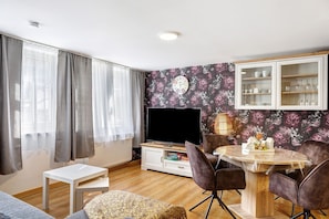 Ein Wohnzimmer mit Flachbildfernseher, einem Esstisch mit Stühlen, einer Couch, einem kleinen weißen Tisch und einer Vitrine.