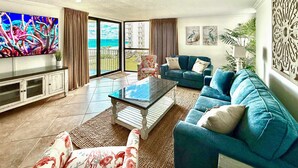 Ein geräumiges Wohnzimmer mit einem großen Fernseher, einem blauen Sofa, einem Couchtisch und Meerblick durch die Schiebeglastüren.