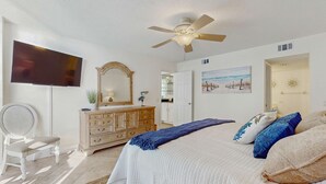 Ein Schlafzimmer mit Bett, Deckenventilator, Fernseher, Kommode, Stuhl und einem Bild an der Wand.