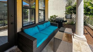 A covered patio with a wicker sofa, a grill, and a white column. - Villa Lago - Tuscany Time (Miramar Beach)