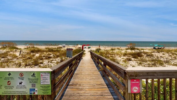 Ein hölzerner Steg führt zu einem Strand mit blauem Himmel und ruhigem Wasser.