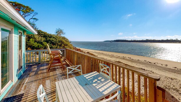 A wooden deck with a table and chairs, overlooking a beach and the sea.