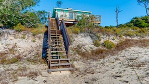 A wooden staircase leading to a turquoise house with a balcony, surrounded by dry vegetation and sand.