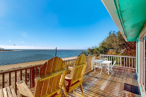 A wooden deck with Adirondack chairs and a table overlooking a beach and ocean.
