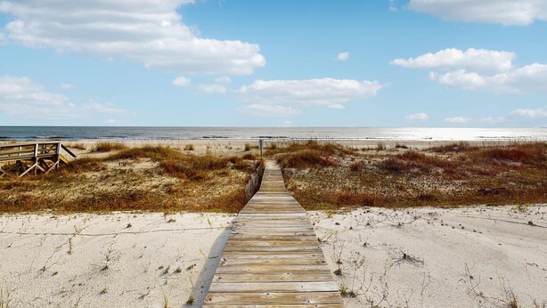 Een houten looppad dat naar een strand leidt, met duinen en een heldere lucht.