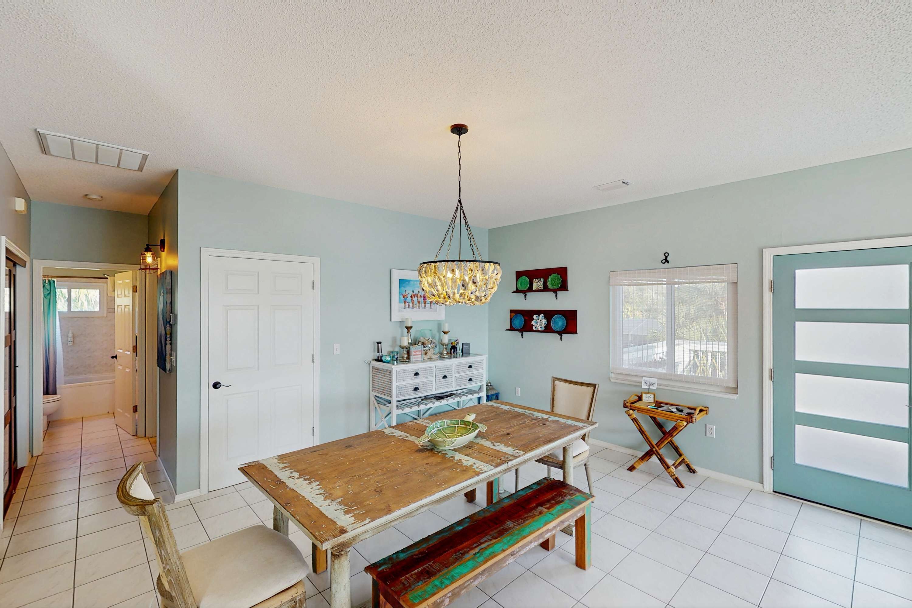 A dining area with a wooden table, bench, and chandelier. There is a door with frosted glass panels and a window with a view.