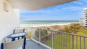 Ein Balkon mit Tisch und Stühlen, der Blick auf einen Strand mit einem hölzernen Steg und ein Gebäude im Hintergrund gewährt.