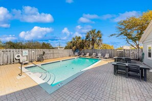Une piscine rectangulaire entourée d’une clôture en bois, avec des chaises longues et une table équipée de bancs.