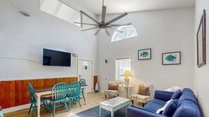 Una sala de estar con ventilador de techo, televisor de pantalla plana, mesa de comedor con sillas, sofá y una mesita de noche con lámpara.
