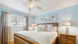 Quarto com cama, mesa de cabeceira de madeira, ventilador de teto e janela com cortinas.