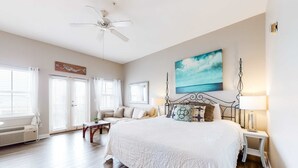 A bedroom with a large bed, a ceiling fan, a painting of the ocean, and a window with curtains. - Cozy Hotel-Style- near beach & dining with kitchenette, pools, hot tub, & tennis (Santa Rosa Beach)