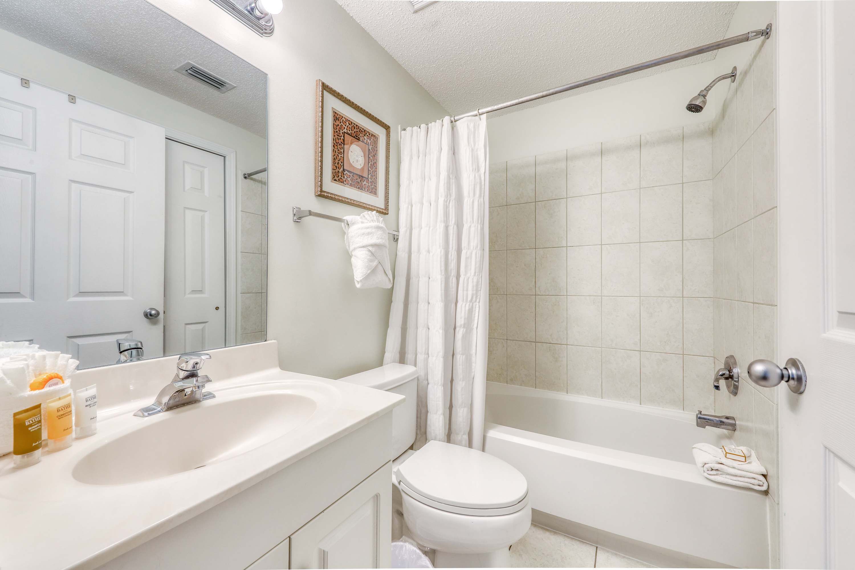 Une salle de bain avec un lavabo, des toilettes et une baignoire, tous de couleur blanche. On y trouve un rideau de douche, un porte-serviettes et un tableau encadré accroché au mur.