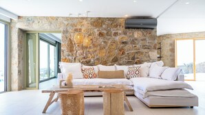 A modern living room with a stone wall, a sectional sofa, a coffee table, and a large window. - Citadel Natural Stone Villa (Mykonos)