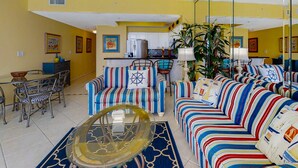 A living room with striped sofas, a glass coffee table, and a dining area with metal chairs. - Majestic Sun A806 Beachfront Views & Pool Fun! (Miramar Beach)
