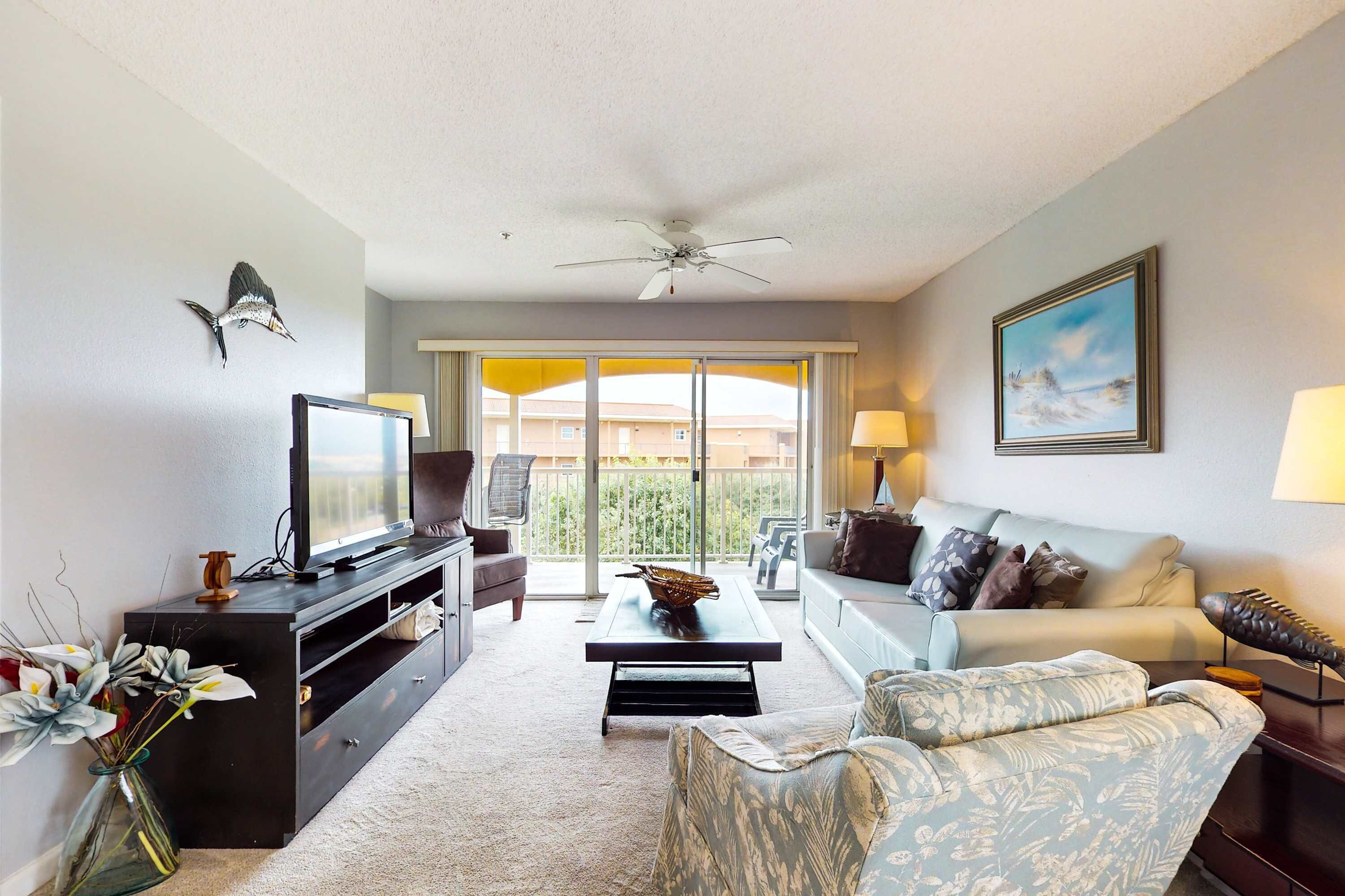 A living room with a flat-screen TV, a ceiling fan, a sofa, a coffee table, and a view of a balcony with outdoor furniture.