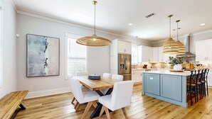 A modern kitchen with a central island, white cabinetry, and a dining area with a wooden table and chairs. - Gorgeous coastal home with shared pools, golf cart, and beach access (Miramar Beach)