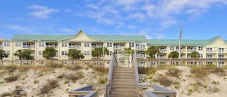 A wooden boardwalk leading to a beachfront hotel with multiple balconies and a green roof.