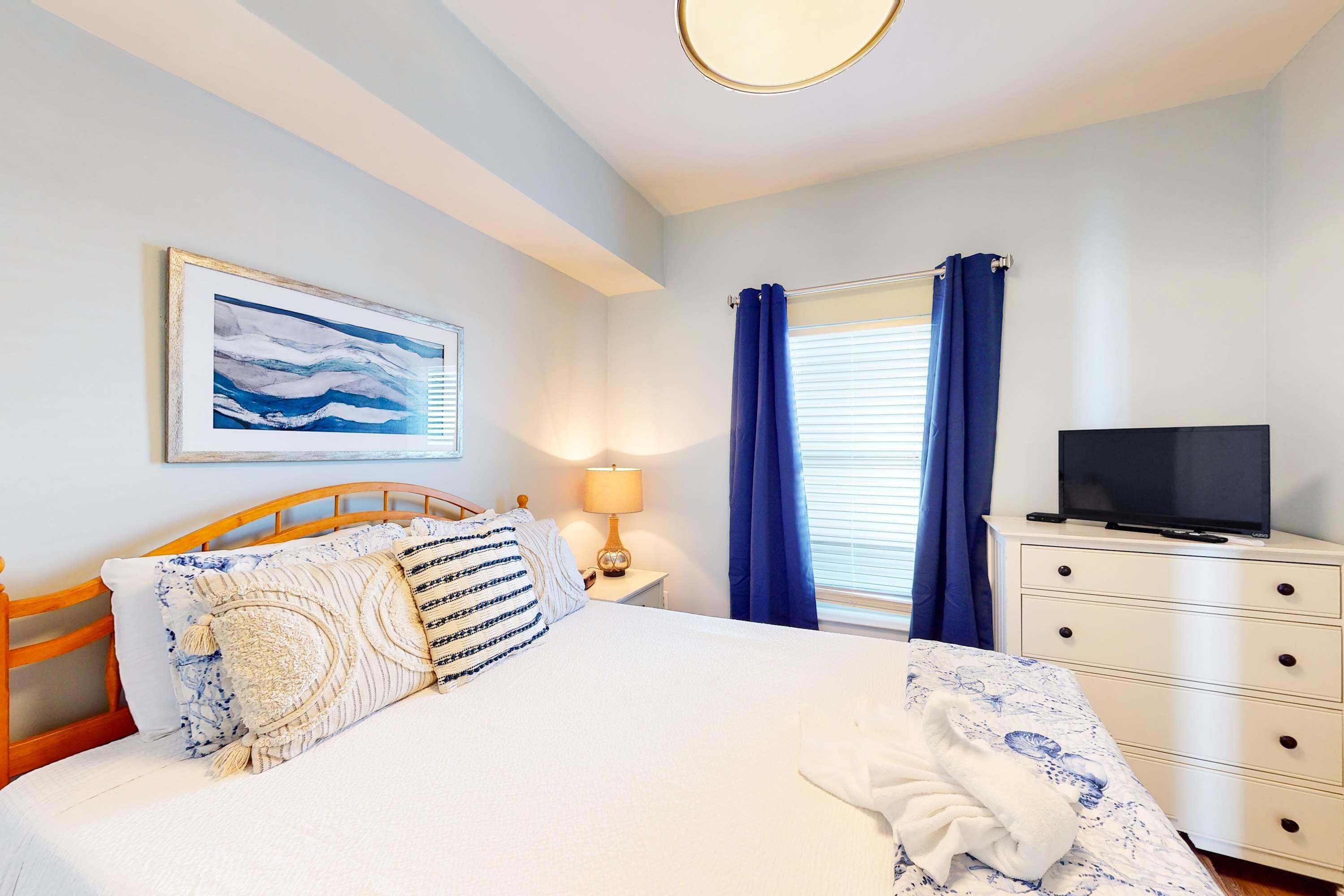 Un dormitorio con cama, televisor sobre una cómoda, cuadro en la pared y ventana con cortinas.