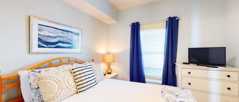 Un dormitorio con cama, televisor sobre una cómoda, cuadro en la pared y ventana con cortinas.