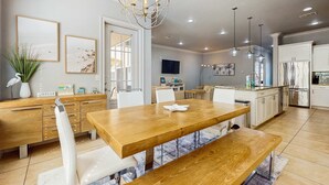 Una cocina moderna con una mesa de comedor de madera, sillas blancas y un banco. Hay lámparas colgantes, un televisor y un refrigerador.