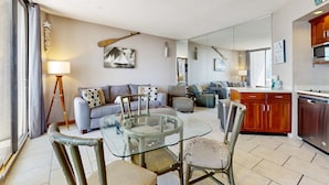 Ein modernes Wohnzimmer mit einem Glastisch, grauen Sofas und einer Küchenzeile mit Holzausstattung und einer Mikrowelle.