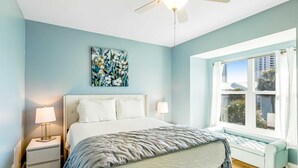 Quarto com cama, abajures, ventoinha no teto, janela com cortinas e um quadro na parede.