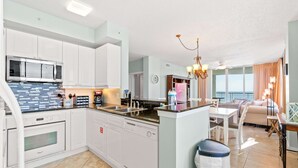 Eine moderne Küche mit weißen Schränken, einem Einbauofen und einem Geschirrspüler. Ein Essbereich mit Tisch und Stühlen bietet Meerblick durch große Fenster.