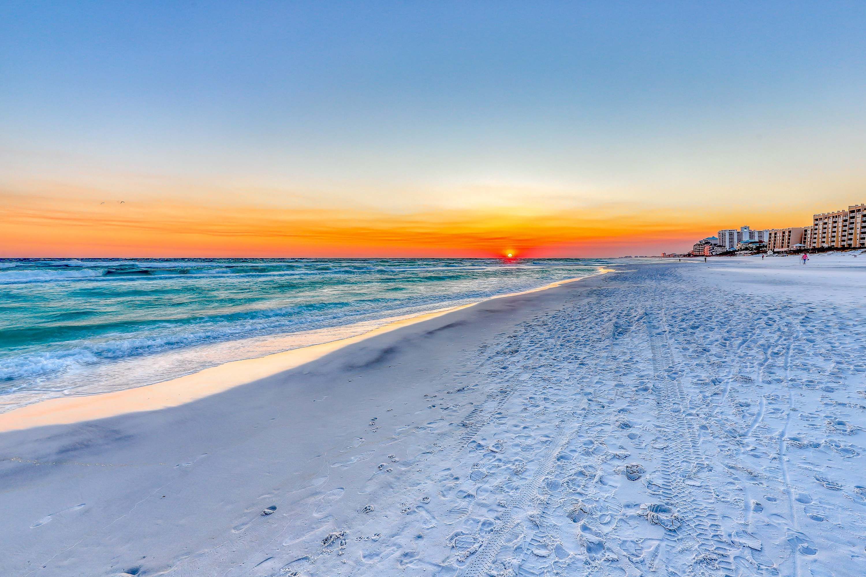 Ein Strand mit Fußabdrücken im Sand, ein Sonnenuntergang im Hintergrund und Gebäude entlang der Küste.