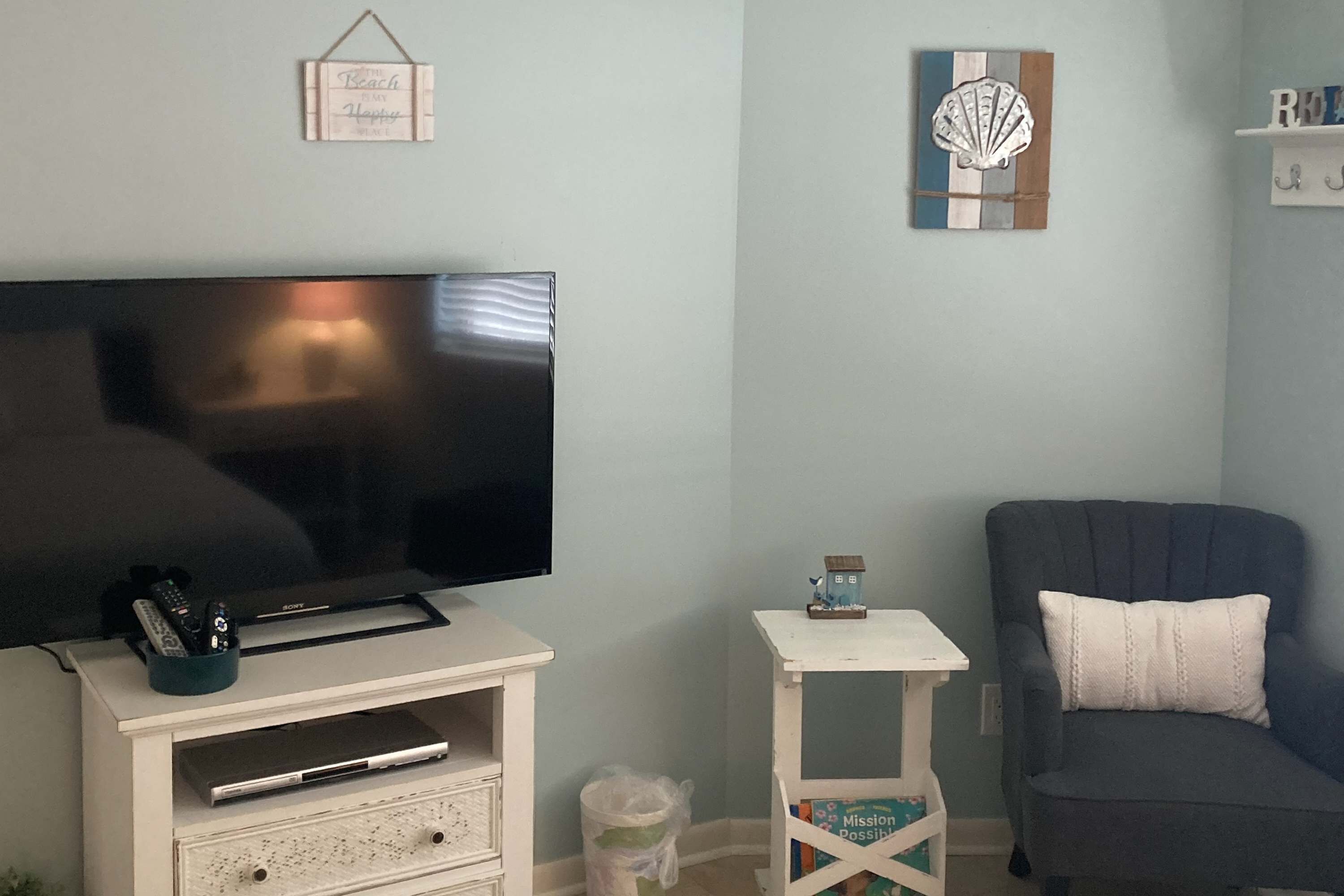 A living room with a flat-screen TV on a white entertainment unit, a small white side table with a decorative item, a dark blue armchair, and a wall-mounted shelf with books.