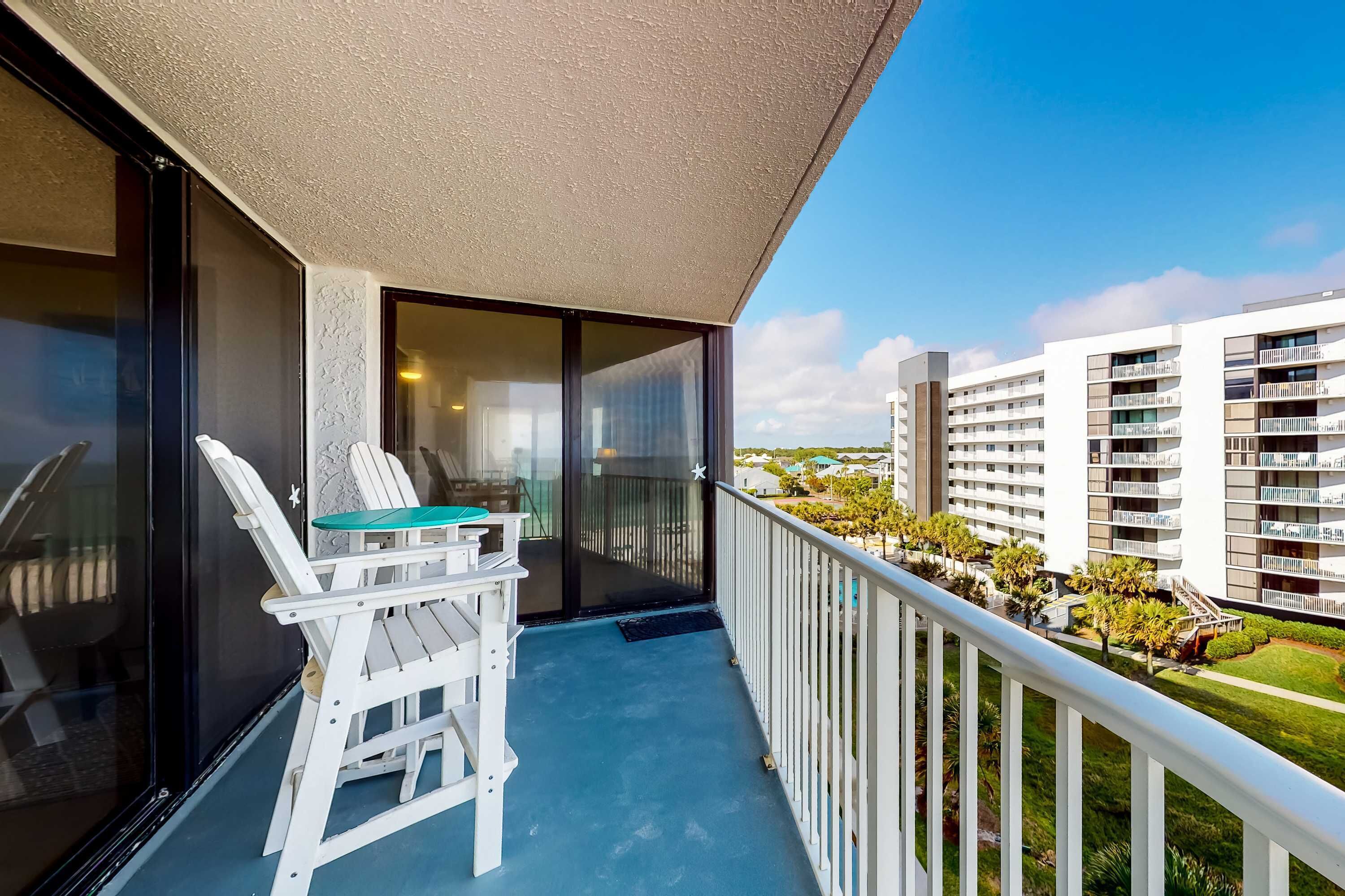 A balcony with a table and chairs, a glass door, and a view of a residential area with buildings and greenery.