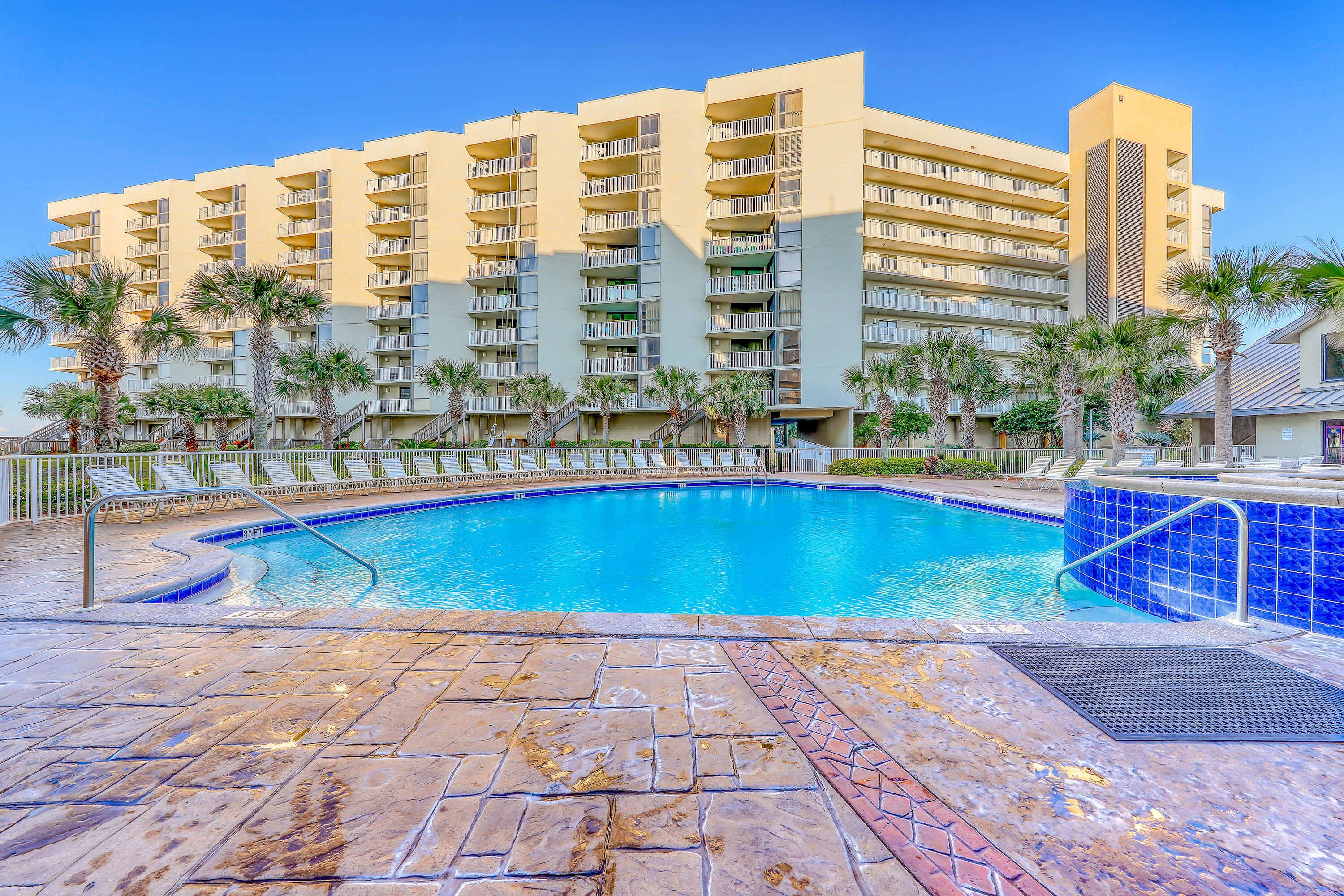 A hotel with a swimming pool, palm trees, and a clear blue sky.