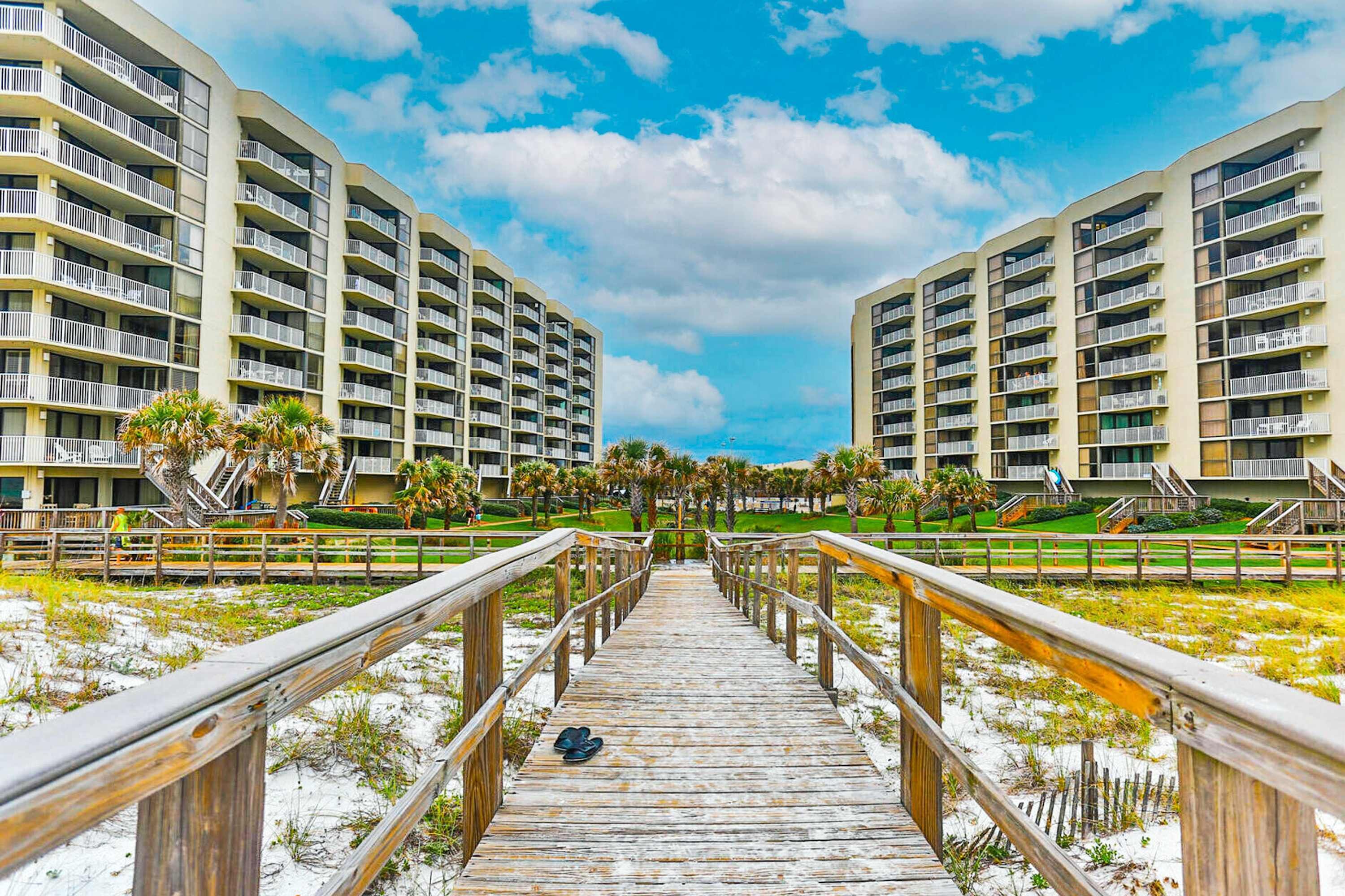 Une promenade en bois mène à un hôtel en bord de mer, doté de nombreux balcons, sous un ciel dégagé.
