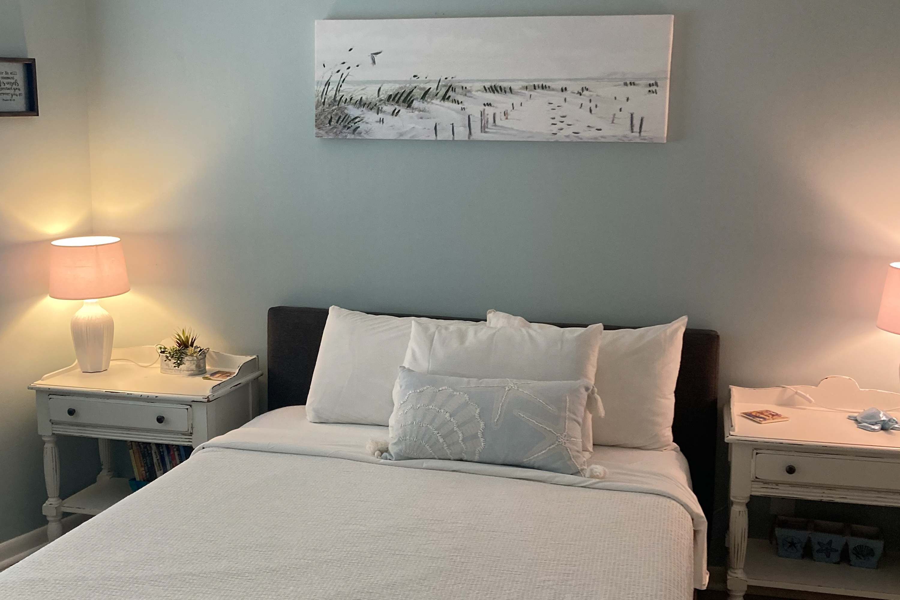 A neatly made bed with white bedding, two bedside tables with lamps, and a beach-themed painting on the wall.