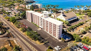 Un hotel de varios pisos con estacionamiento, situado cerca de la costa, con palmeras y vistas despejadas al océano.