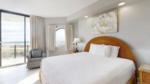 A hotel room with a large bed, a wicker chair, a round window, and a view of the landscape. - Surfside Resort  1211 (3) (Miramar Beach)