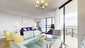 A modern living room with a glass coffee table, comfortable seating, a chandelier, and a large window with a view. - Surfside Resort  1211 (3) (Miramar Beach)