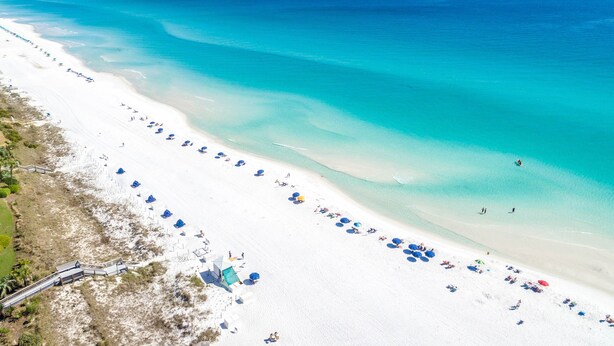 Vista aérea de uma praia com areia branca, águas turquesa cristalinas e guarda-sóis azuis.
