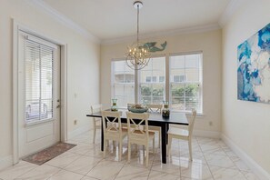 A dining area with a black table, white chairs, a chandelier, and a large window with blinds. - Townhouse with private patio, pool & private beach access - snowbirds welcome (Miramar Beach)