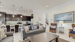 A modern living room with a grey sofa, a wooden coffee table, wicker chairs, and a kitchen in the background.