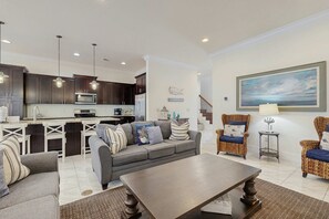 A modern living room with a grey sofa, a wooden coffee table, wicker chairs, and a kitchen in the background. - Townhouse with private patio, pool & private beach access - snowbirds welcome (Miramar Beach)
