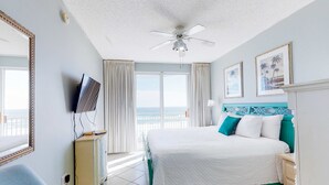 Ein Hotelzimmer mit Bett, Fernseher, Deckenventilator, Fenster mit Vorhängen und Blick auf den Ozean.