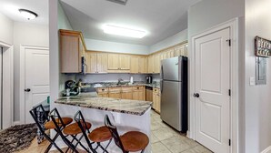Uma cozinha com armários de madeira, uma geladeira de aço inoxidável, uma ilha com bancada de mármore e banquetas, e piso revestido de azulejos.